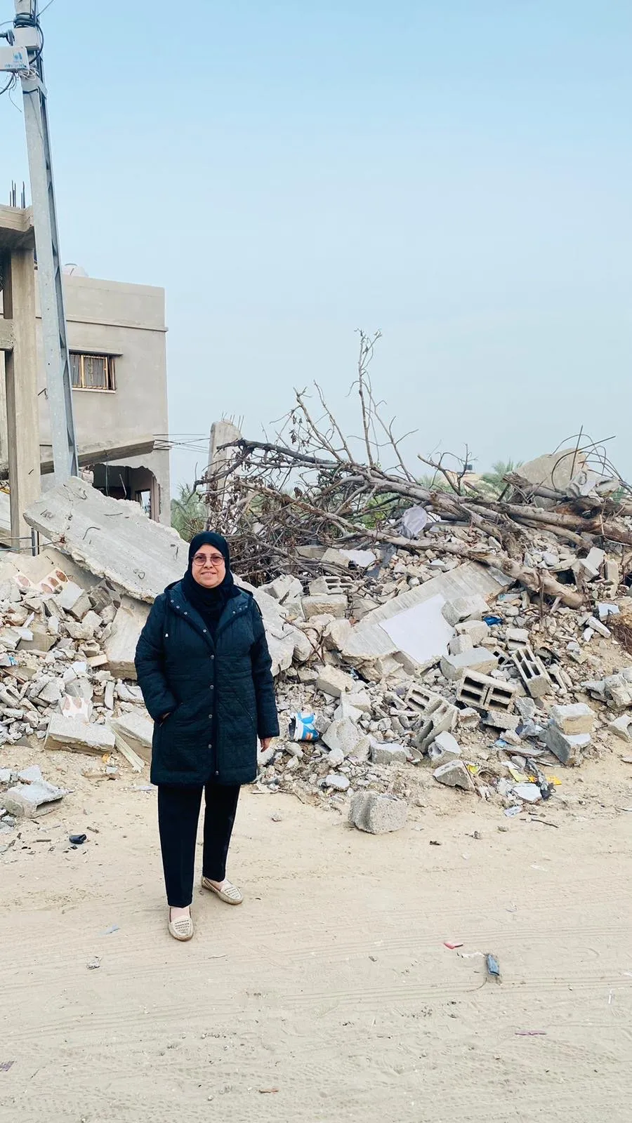 Woman in head covering and glasses stands outdoors in front of a pile of rubble
