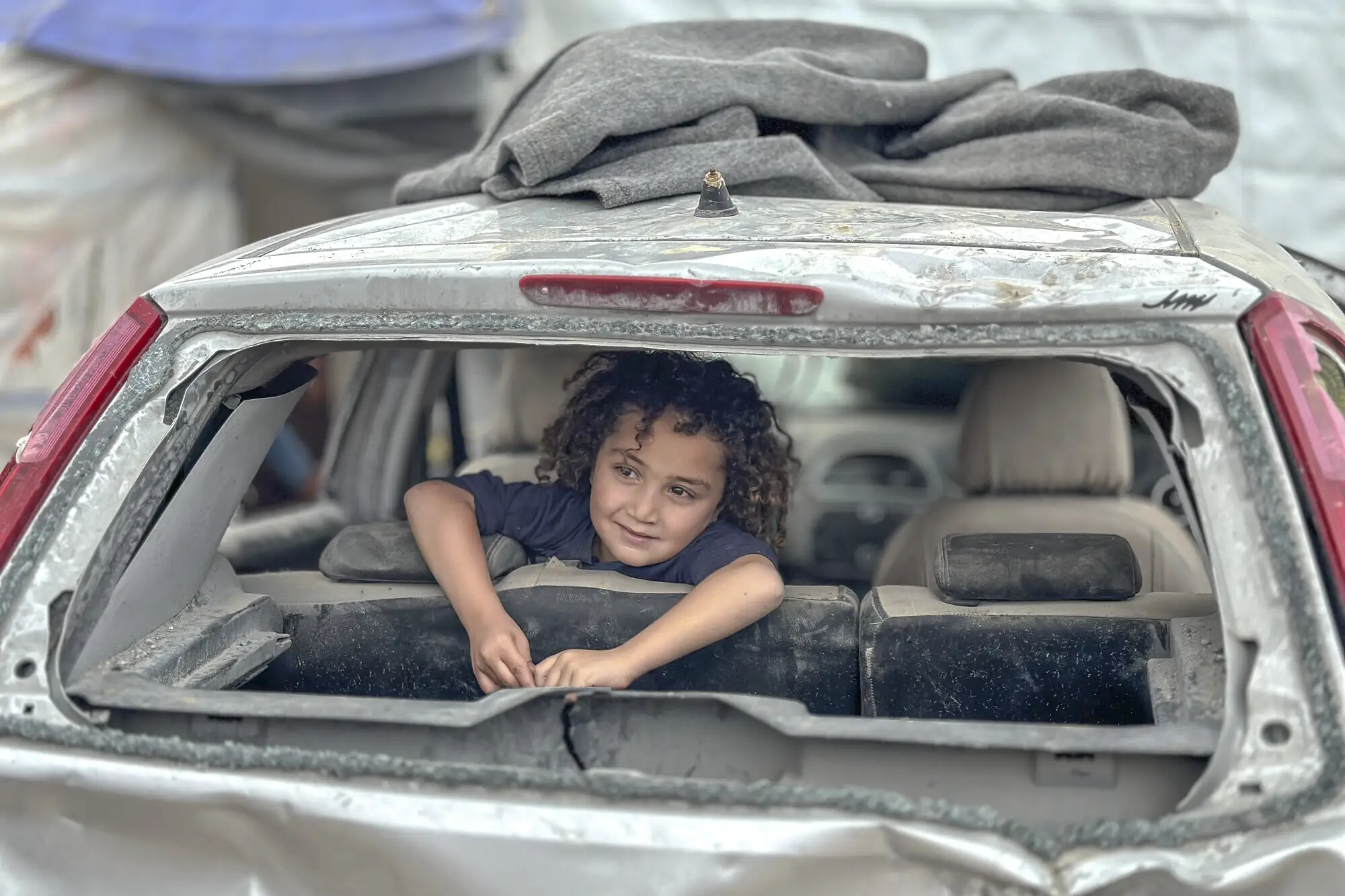 Criança com meio sorriso olha pela janela traseira de um carro danificado