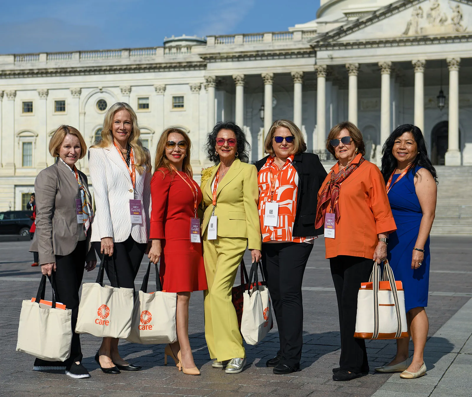 A group of women, many of whom are wearing orange or carrying CARE tote bags, stand together in front of the U.S. Capitol Building in Washington, DC.