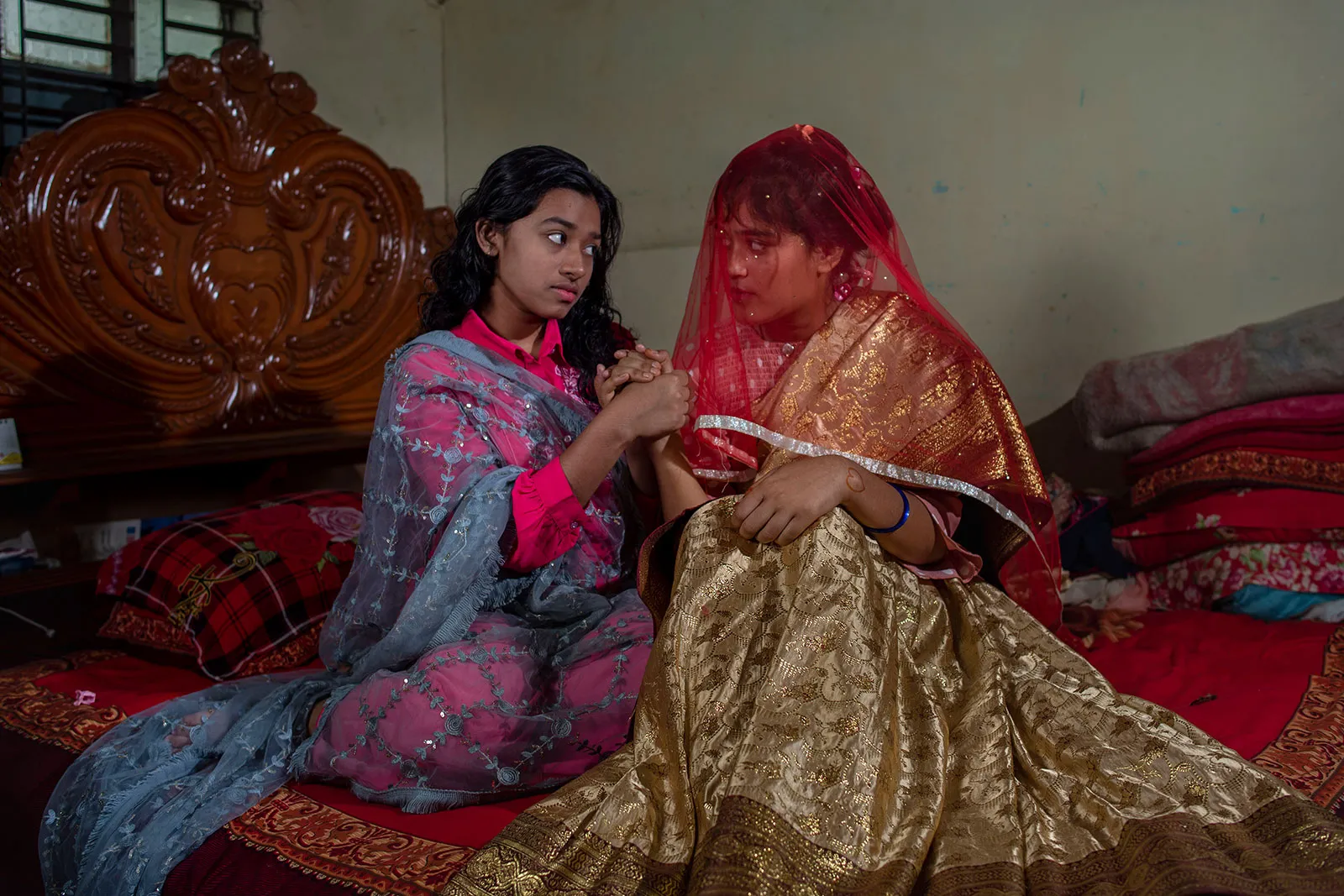 Two teenage Bangladeshi girls sit on a bed with a bright red quilt and clasp hands while facing each other. One of the girls is wearing a red veiled head covering.