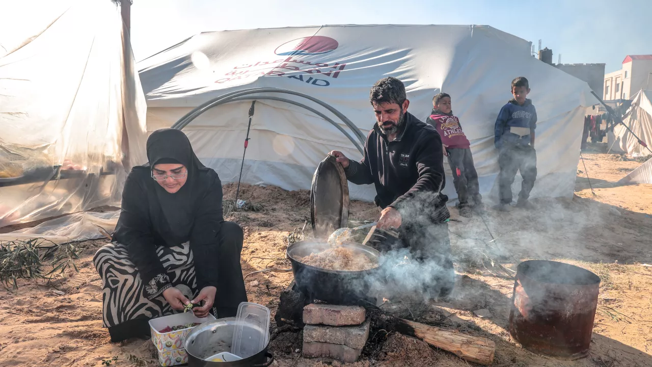 A woman cooks outside of a tent while a man stands behind her in Gaza.