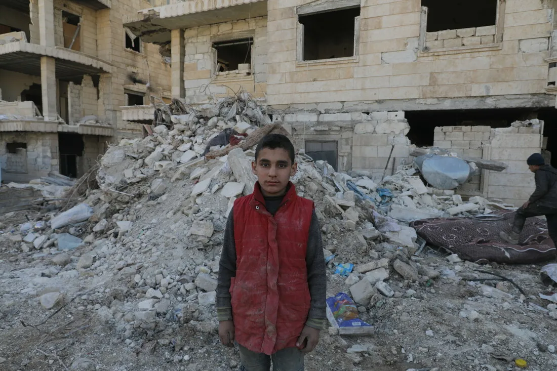 A young boy wearing a red vest stands in front of rubble.