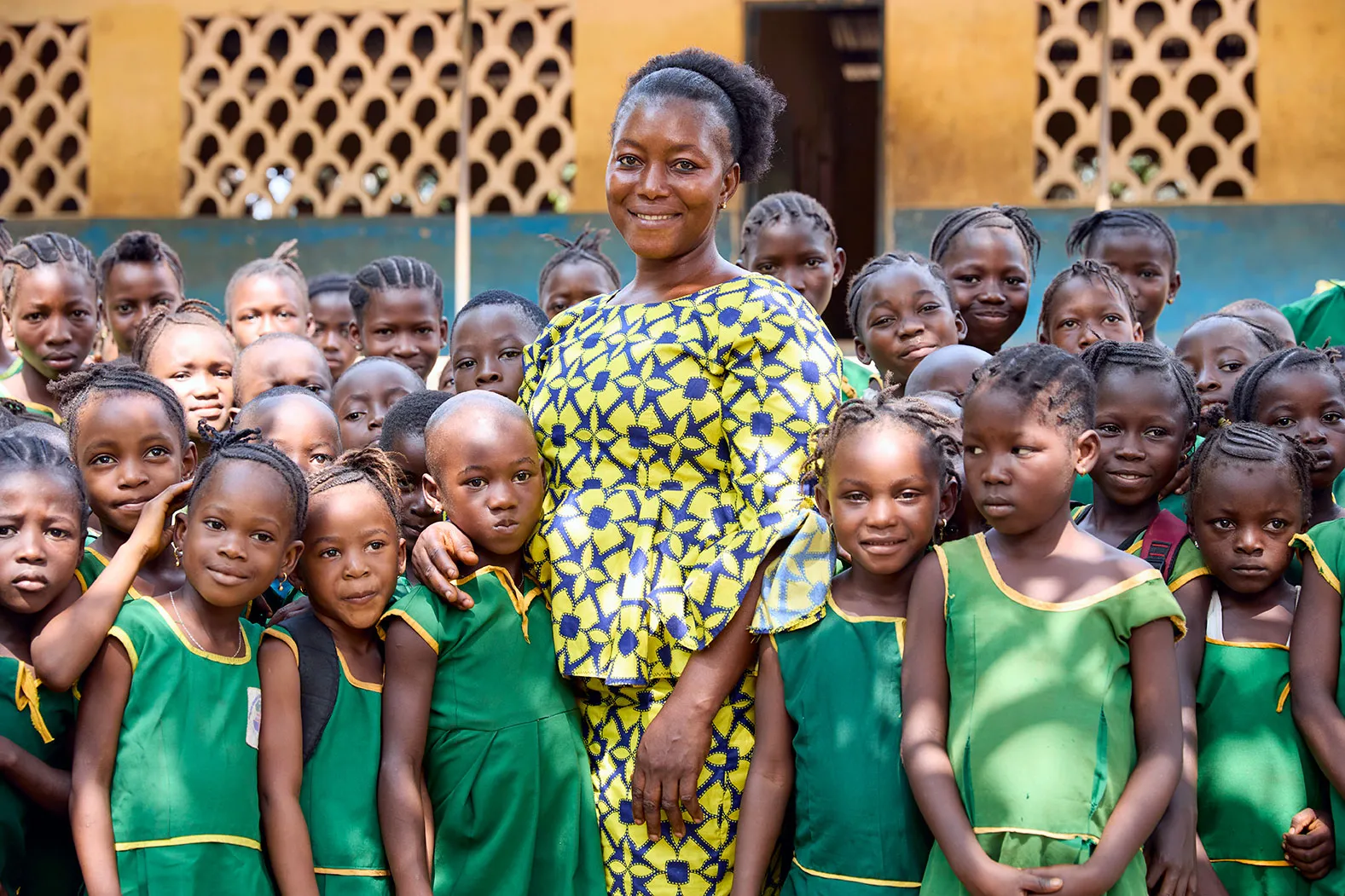 A woman wearing bright yellow is surrounded by children wearing green school uniforms.