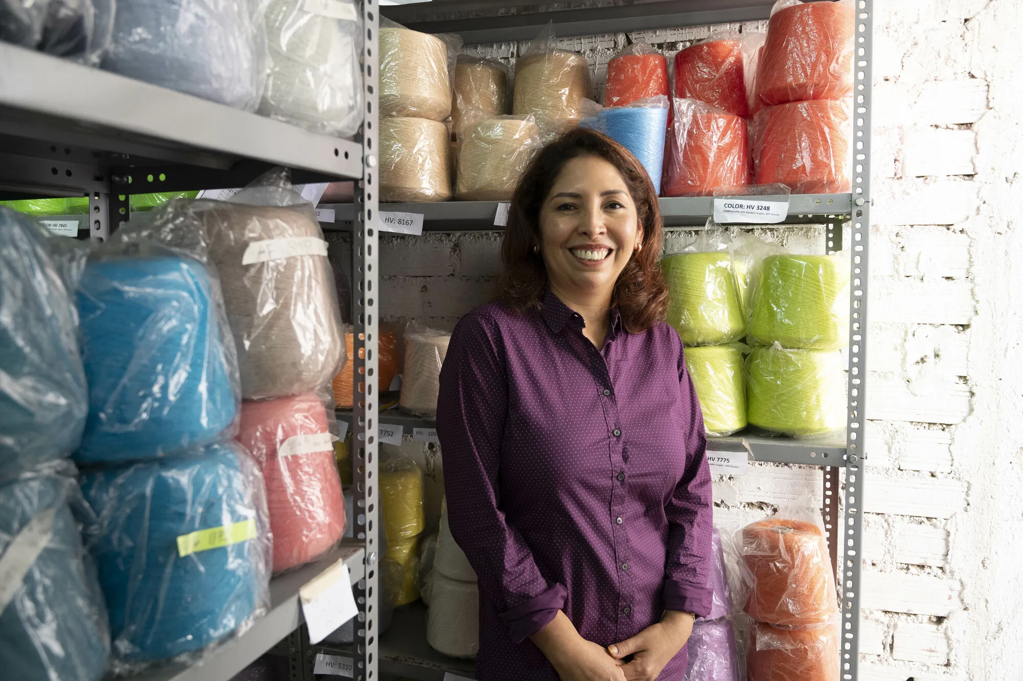 Portrait of a smiling woman wearing a purple shirt, surrounded by spools of yarn and thread