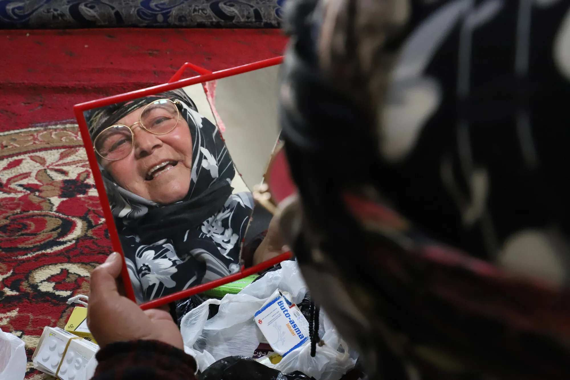 An internally displaced Syrian woman in glasses holding a mirror and gazing at her own reflection with some medicines on her lap.