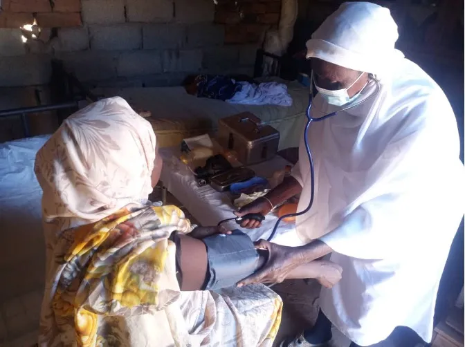One woman in white, head covered covering using a blood-pressure sleeve on another woman whose head is also covered.