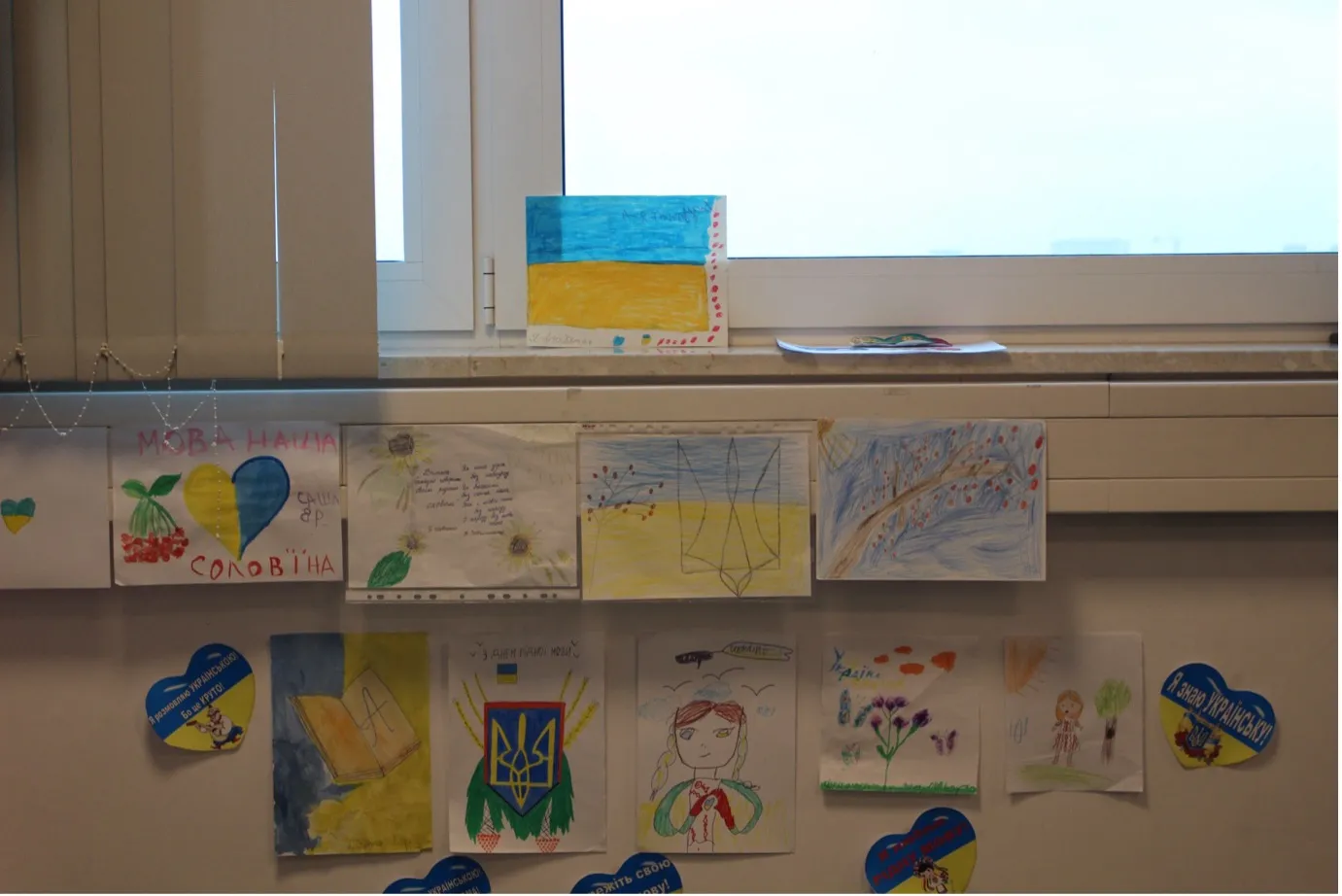 A group of children’s drawings affixed to a wall underneath a window.