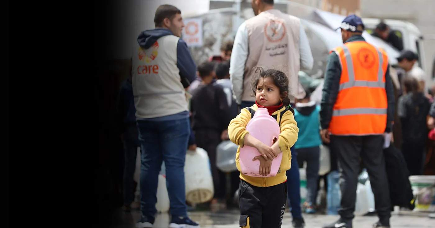 A young girl holds a pink jug close to her chest. In the background are emergency aid workers, including a man in a CARE vest.