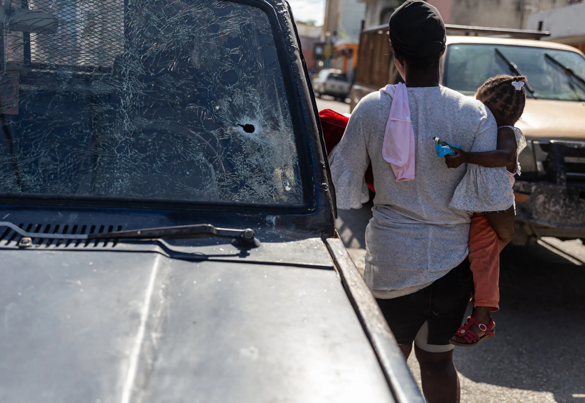 A woman holds a child outdoors, back to camera, next to a car with a bullet hole in the windshield.