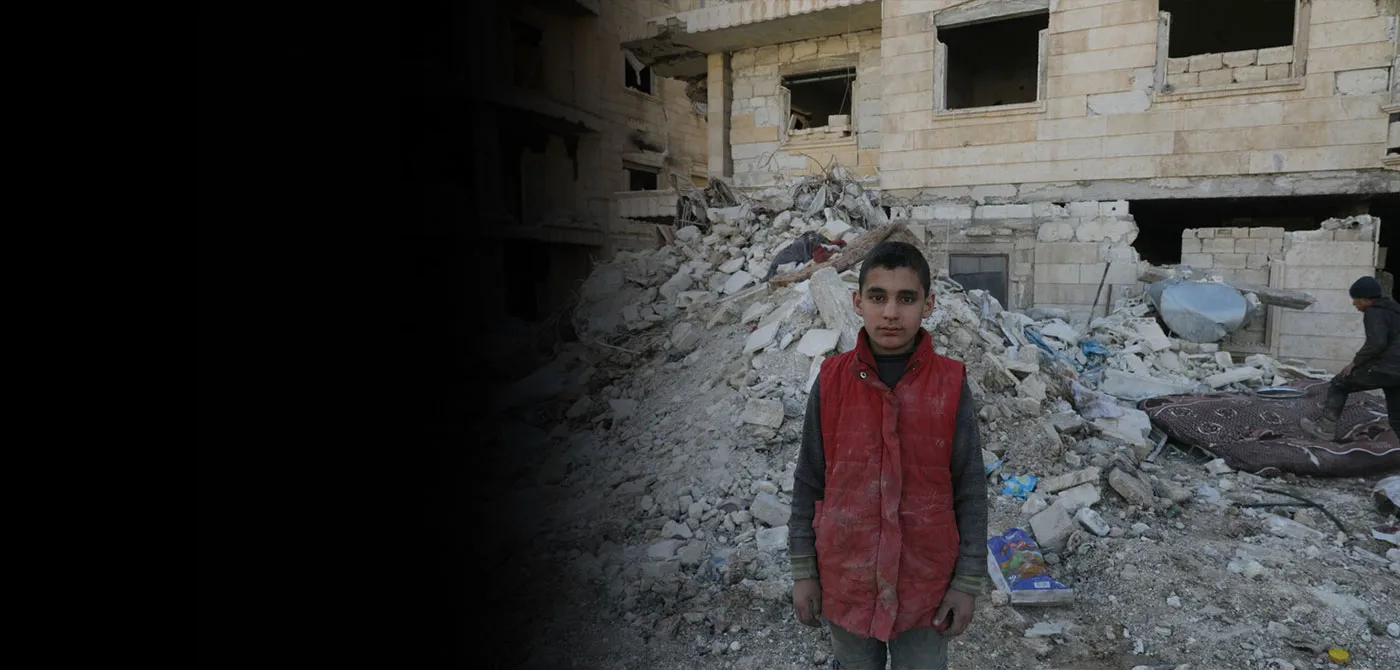A young boy wearing a red vest stands in front of rubble.