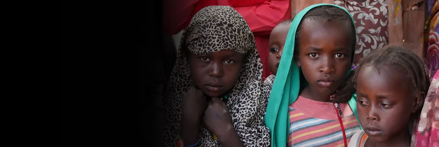 Three Sudanese children stand next to each other, one of them clutching fabric around their head.