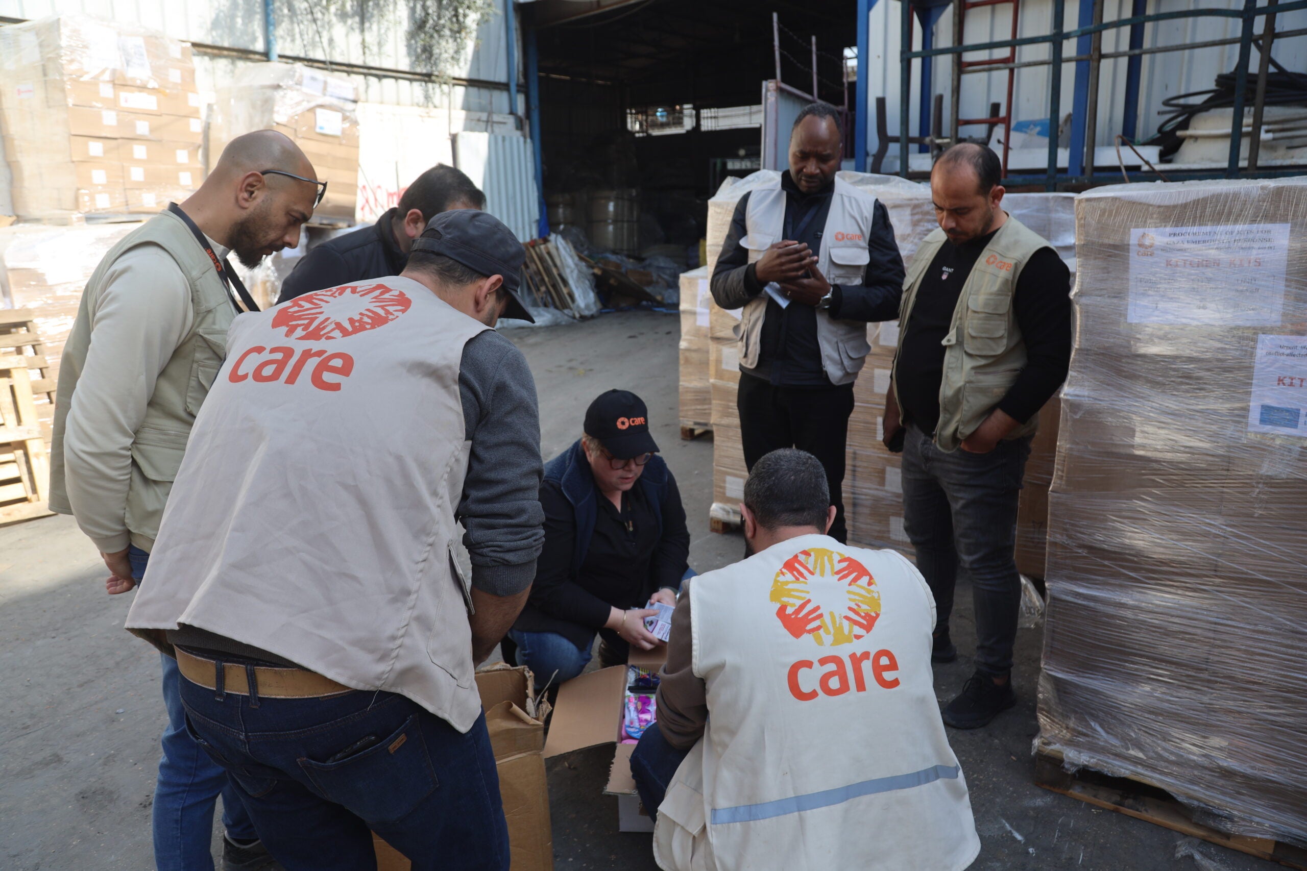 A group of people, several wearing CARE vests, crouch around packaged supplies in a warehouse in Gaza city.