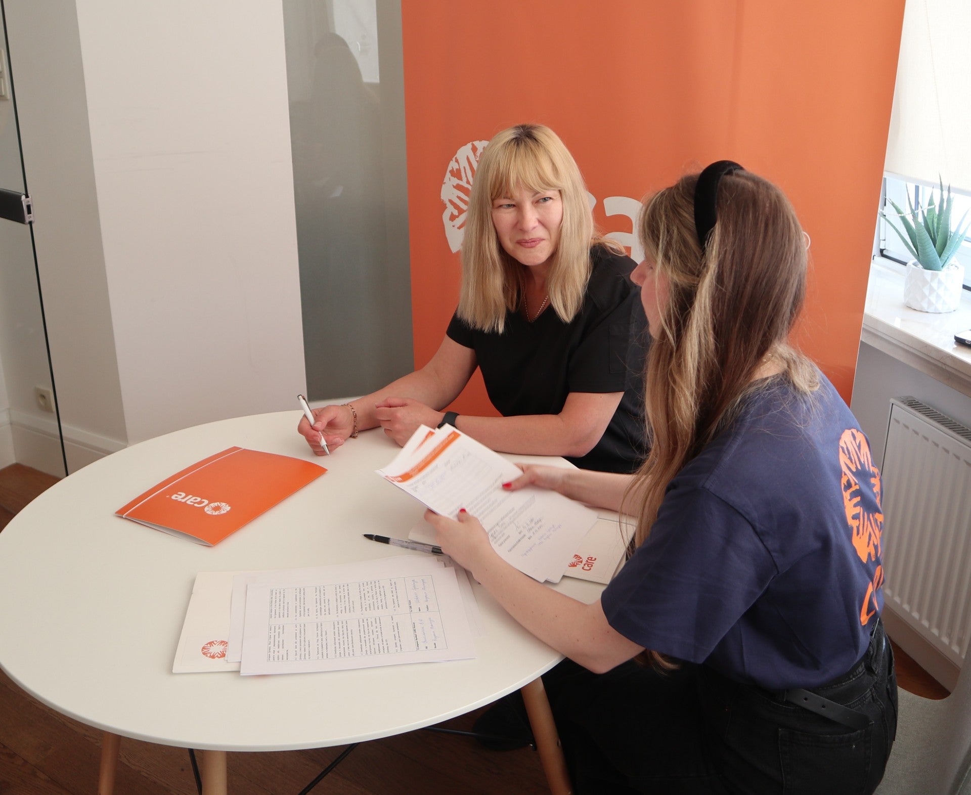 Two women are seated close together at a table, engaged in a discussion. The woman on the left smiles as she looks at the other woman. Several publications featuring the CARE logo are visible on the table, and the CARE logo is also visible on the back of one of the women's t-shirts.