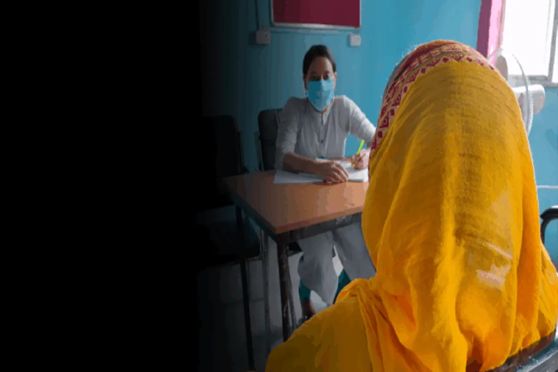 A woman wearing bright yellow clothing is faced away from the camera so her face is not visible. Across from her is a woman at a desk wearing a facemask.