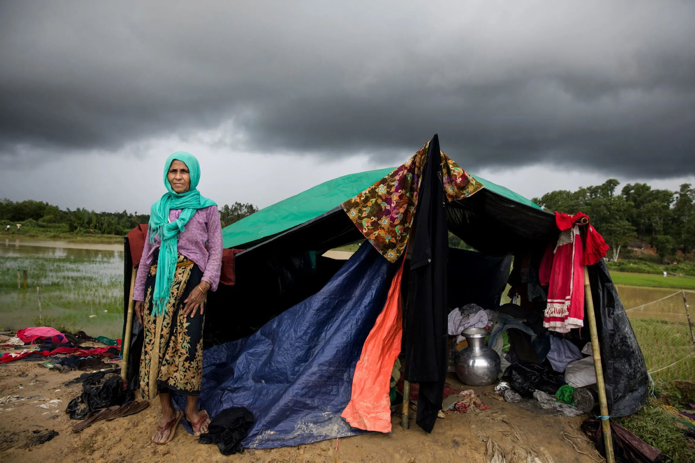 A medium-shot color photo a Rohingya woman standing in front of a makeshift tent with a dark, cloudy sky overhead, a flooded field behind her, and clothes hanging on the side of the tent.
