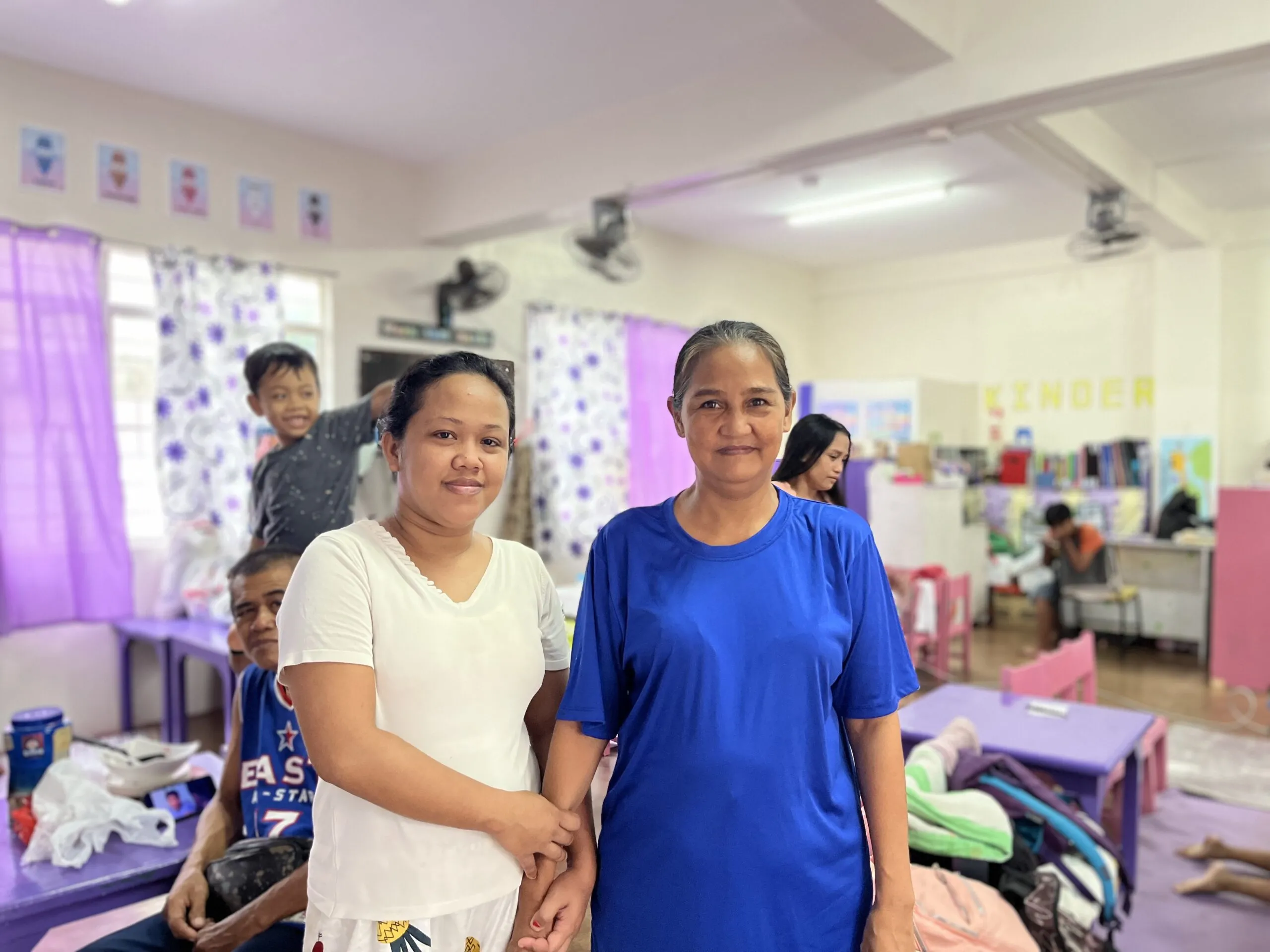 Two women stand in front of their families in a makeshift evacuation center.