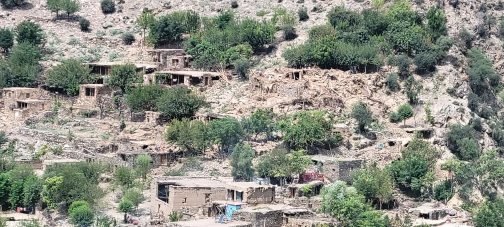 Aerial view of damaged mud-brick homes scattered across hillside terrain in Afghan village after earthquake.