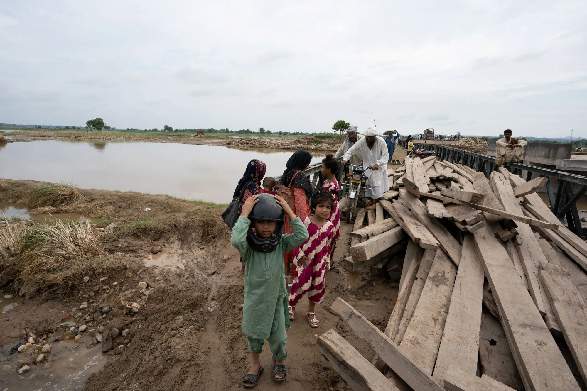 A group of Pakistanis, including children, walk a muddy path beside floodwaters and a damaged bridge. A boy in a helmet is in the foreground as a man in traditional white clothing pushes a motorcycle loaded with people and belongings across the bridge.