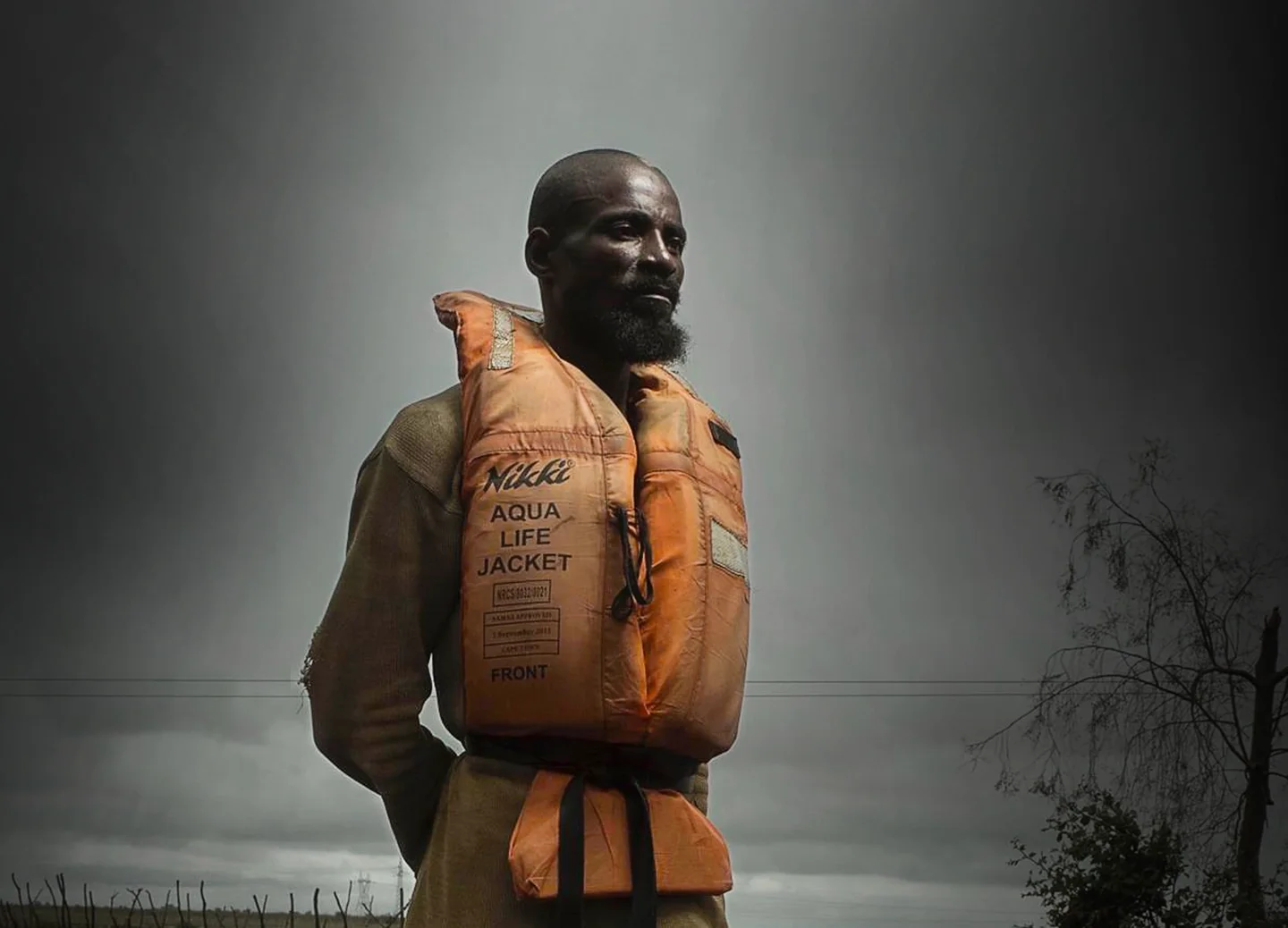 A man with a beard stands outdoors, wearing an orange life jacket.