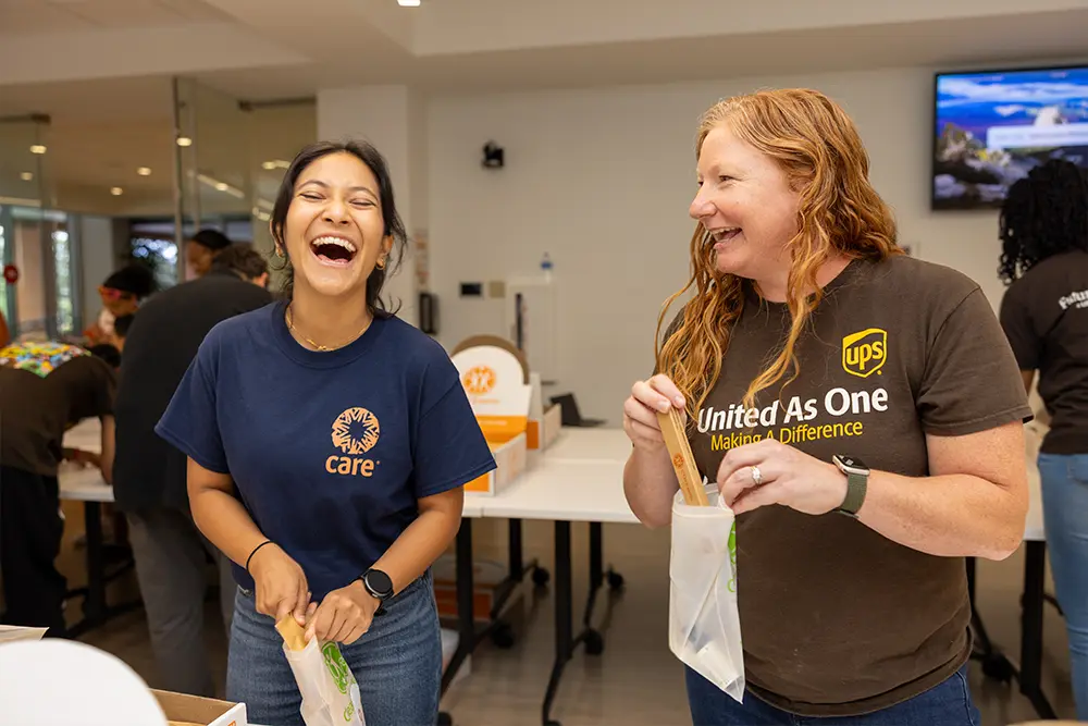Two women, one wearing a CARE t-shirt and one wearing a UPS t-shirt, laugh while packing bags.