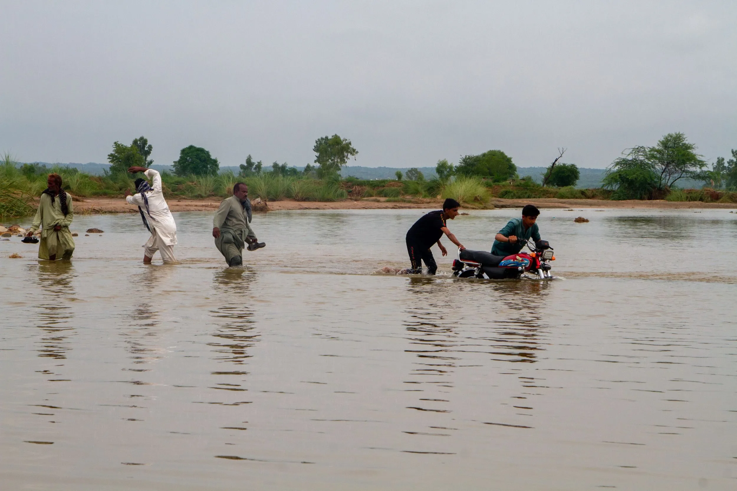 A group of Pakistani men wade through floodwater, pushing a motorcycle across a submerged roadway. One man lifts his white pants to keep them dry, while others carefully move the vehicle, with trees and brush visible on the distant shore under a cloudy sky.