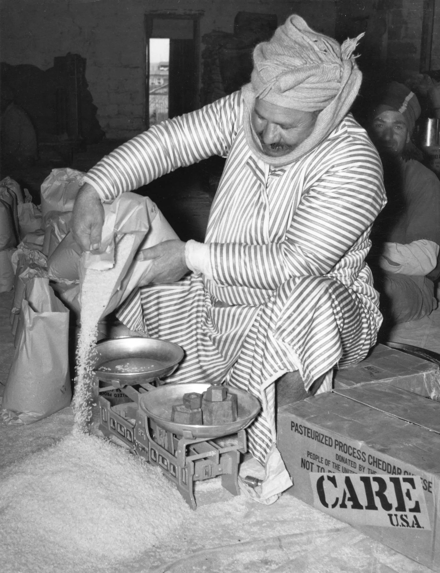 A historic black and white photo shows a man, in traditional Arabian robes and a turban weighing rice on a scale, with rice spilled on the floor around him. A box labeled 