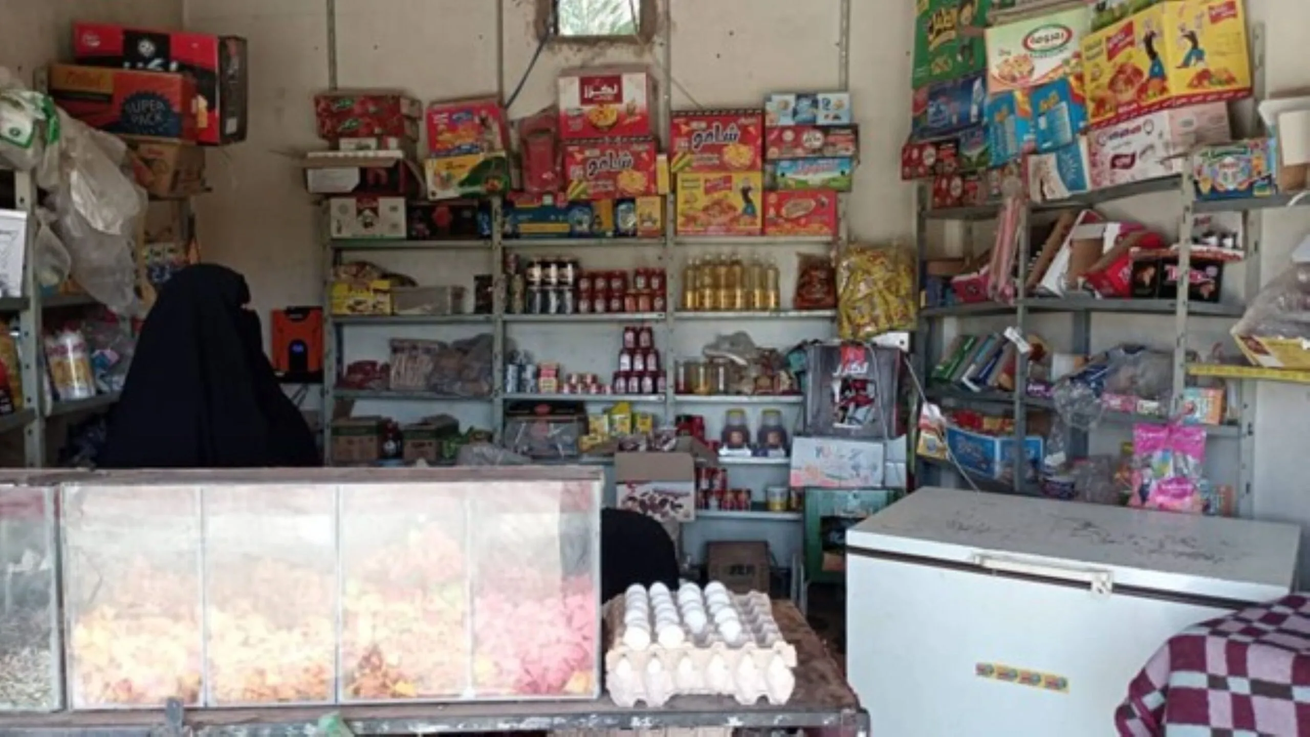An interior shot of a small, well-stocked grocery shop or kiosk in Syria, with the woman owner, a member of Village Savings and Loan Association or VSLA, seated behind the counter wearing a black niqab or veil.