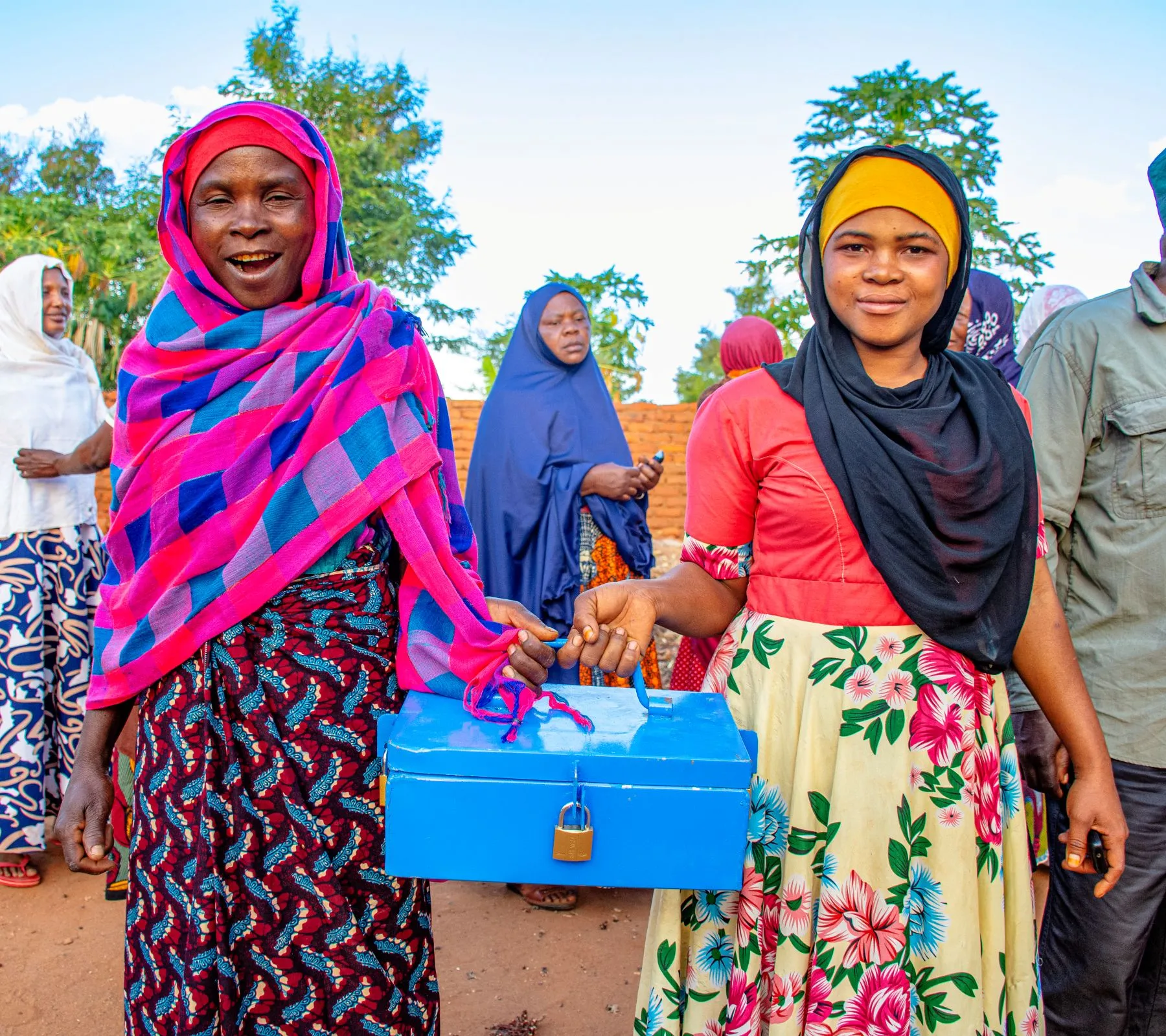 Two Tanzanian women, who are VSLA group members, proudly hold their locked bright blue metal savings box outdoors. The woman on the left, wearing a pink and blue shawl, smiles broadly, highlighting the importance of the communal funds they manage.