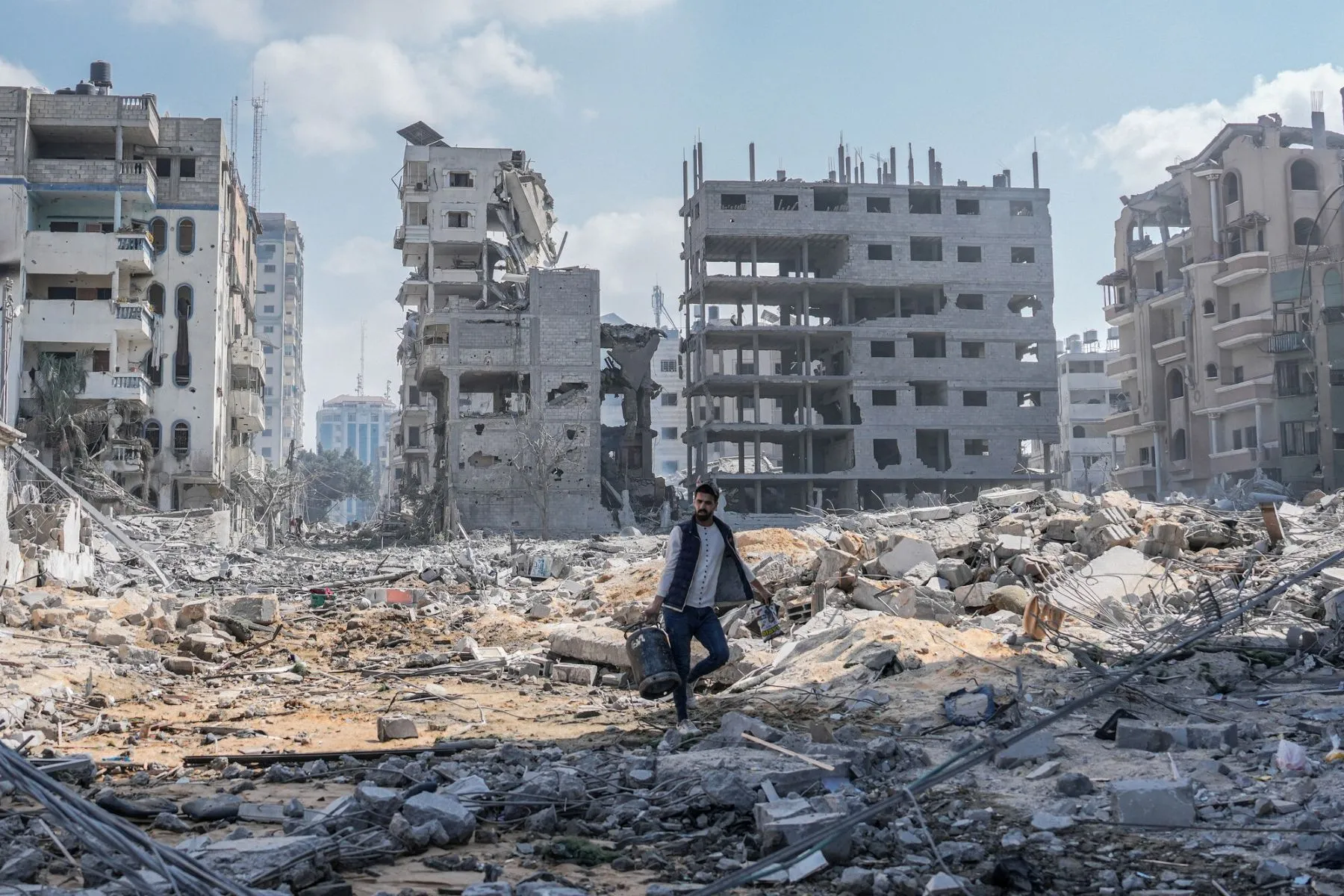 A man in Gaza walks alone through a vast landscape of rubble and debris, carrying a cooking gas cylinder. Behind him stand multiple apartment buildings that have been severely damaged or completely destroyed by strikes.