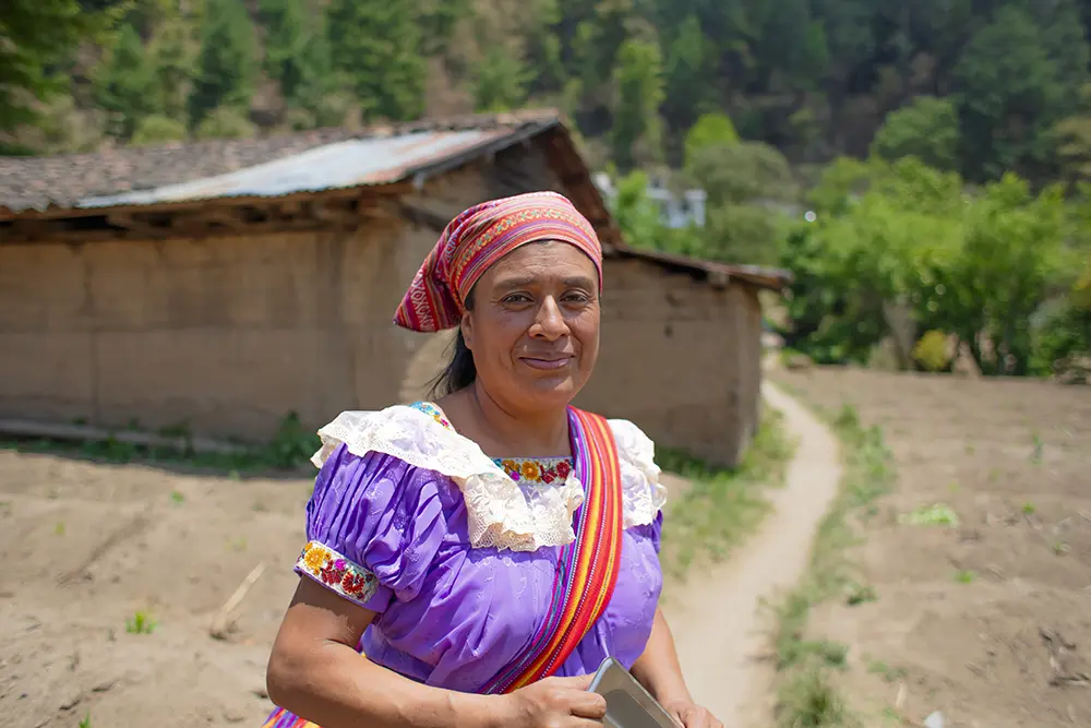 A Guatemalan woman with a bag slung over her shoulder stands on a path outside a small home.