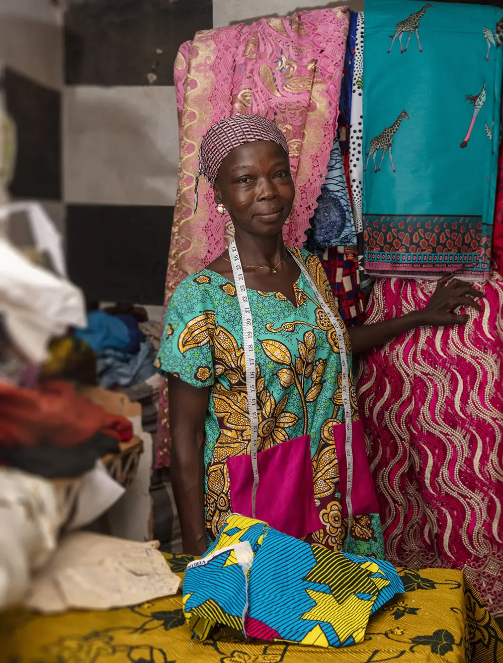 A woman with a tape measure hanging around her neck stands in front of cloths with vibrant patterns.