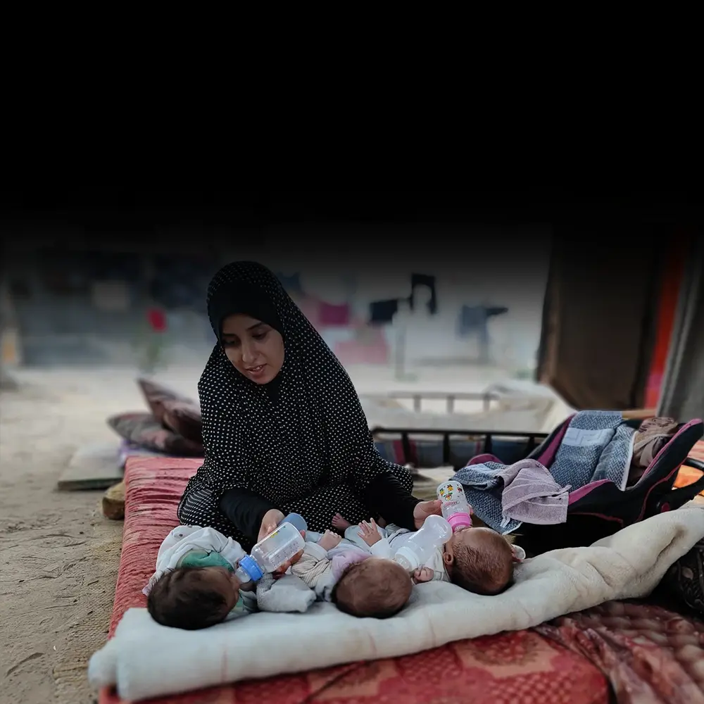 A mother in Gaza feeds two of her triplet babies, holding two bottles in her hands.