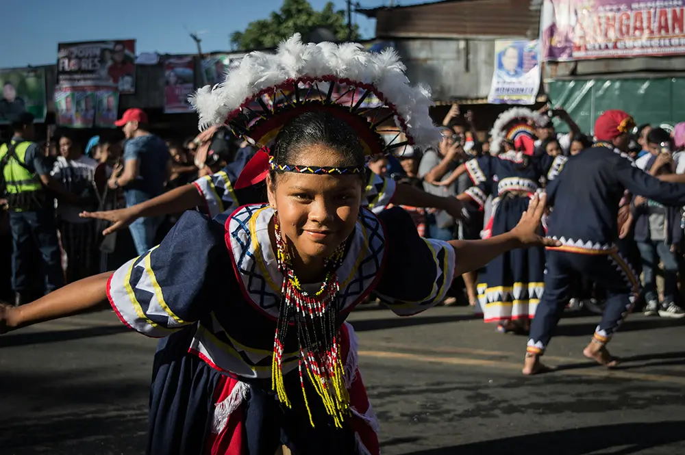 An indigenous girl in traditional wear bends down and smiles while dancing.