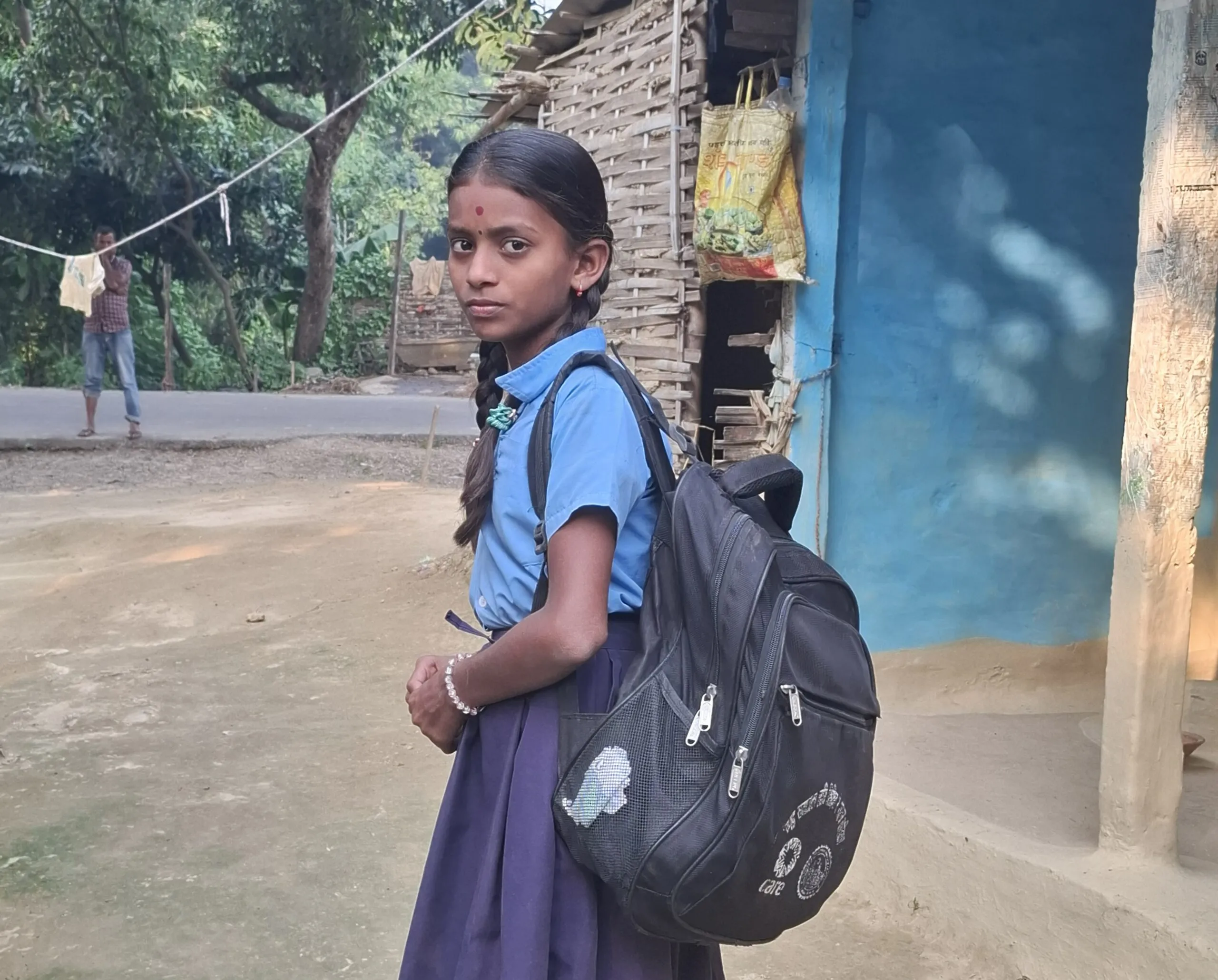 Adolescent Nepalese girl in school uniform with a large black backpack, standing outside a rustic home.