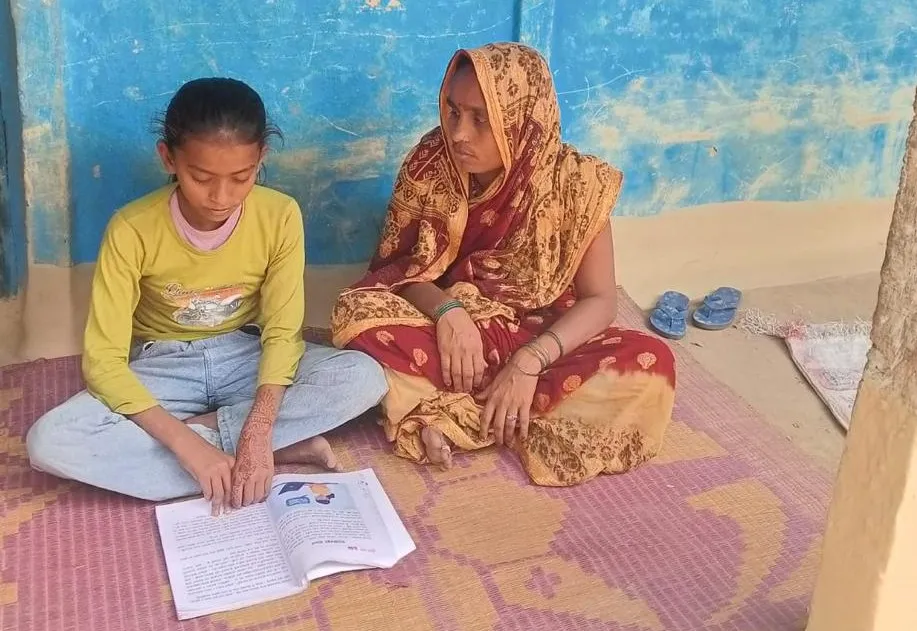 Radhika and her mother, Sundar Kala, sitting on a mat outside a blue house, with Radhika reading from an open textbook.