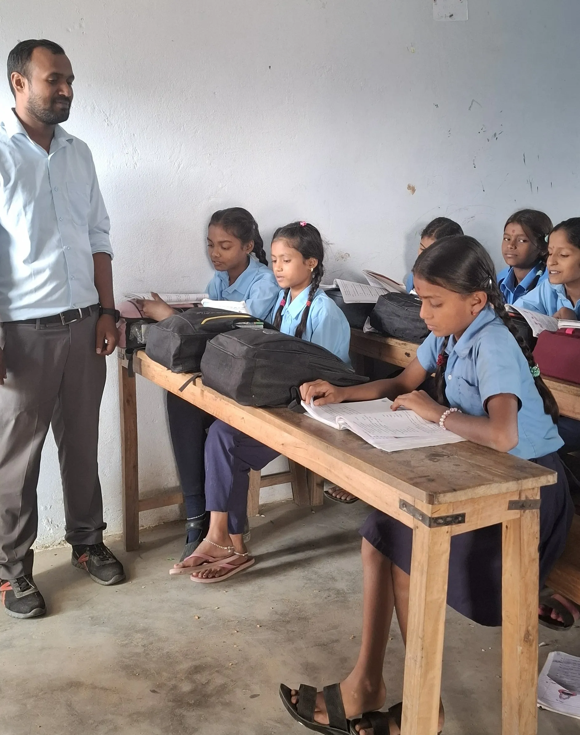 A male teacher stands in a basic classroom watching several schoolgirls in blue uniforms sitting at wooden desks and writing in their notebooks.