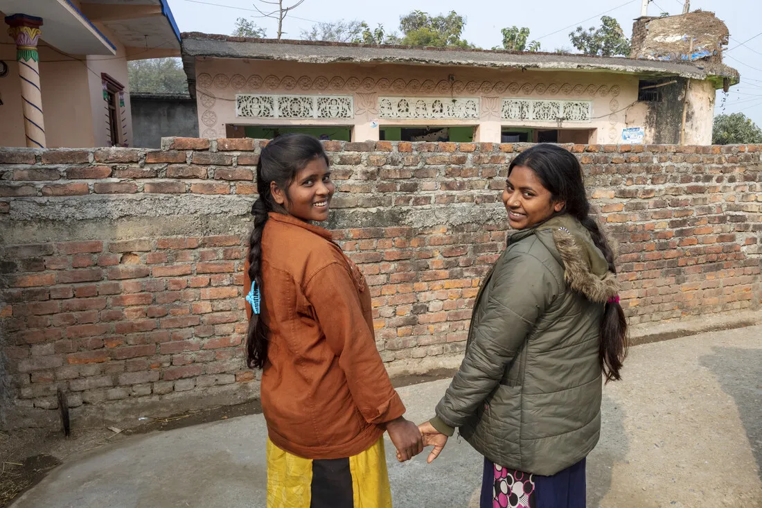Two young Nepalese girls, smiling and holding hands, stand in front of a weathered brick wall and traditional village buildings.