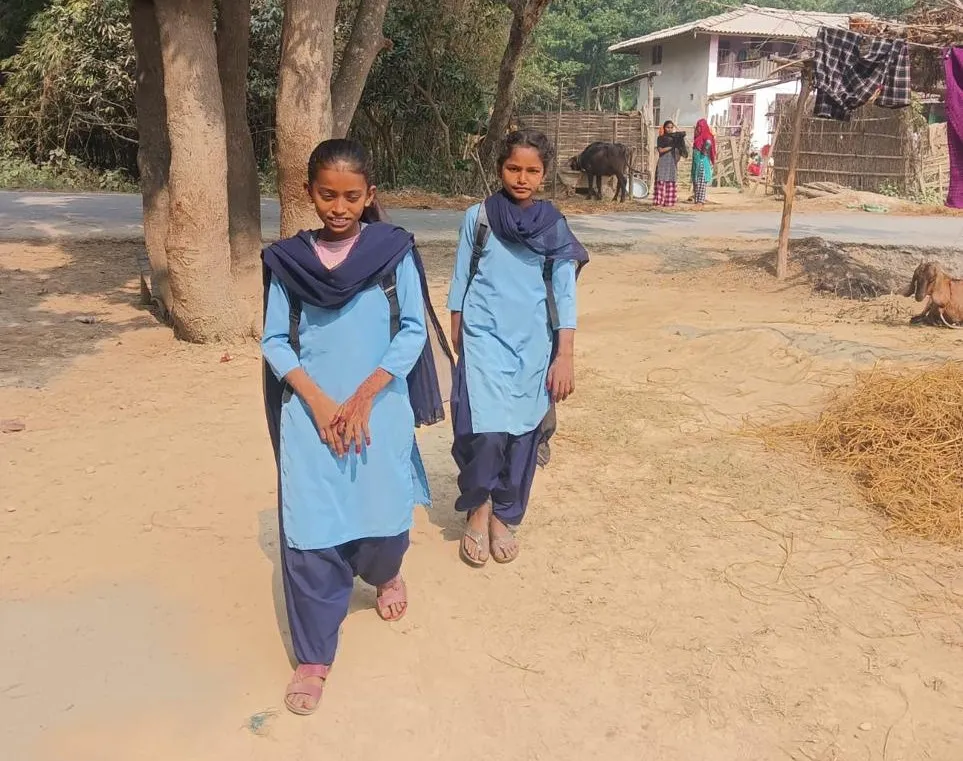 Two schoolgirls, including Radhika, wearing blue uniforms and carrying backpacks, walk along a dusty village road.