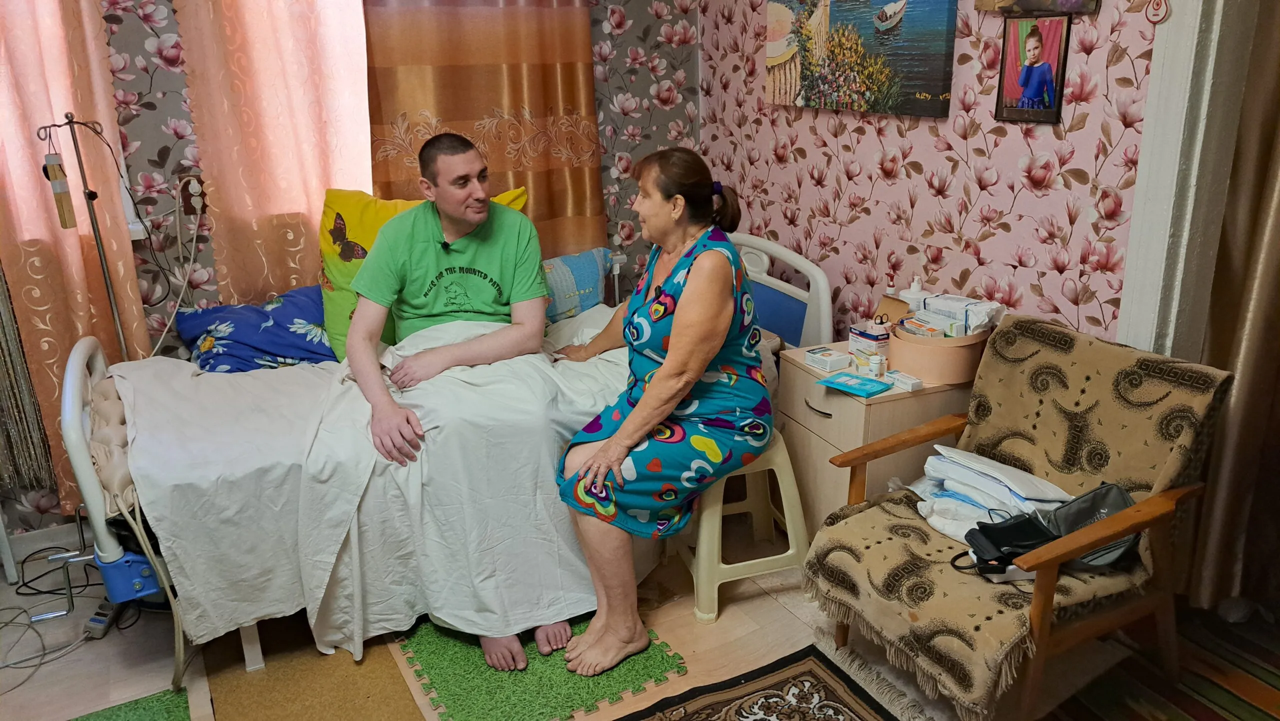 A mother sits with her adult son, who is in a medical bed.
