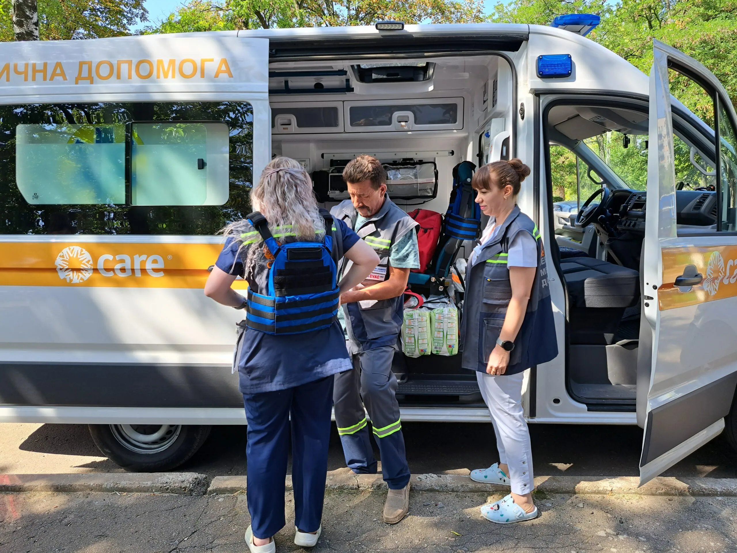 Three people in high-vis gear stand outside a CARE-branded ambulance. One helps the other put on a bright blue bullet proof vest.