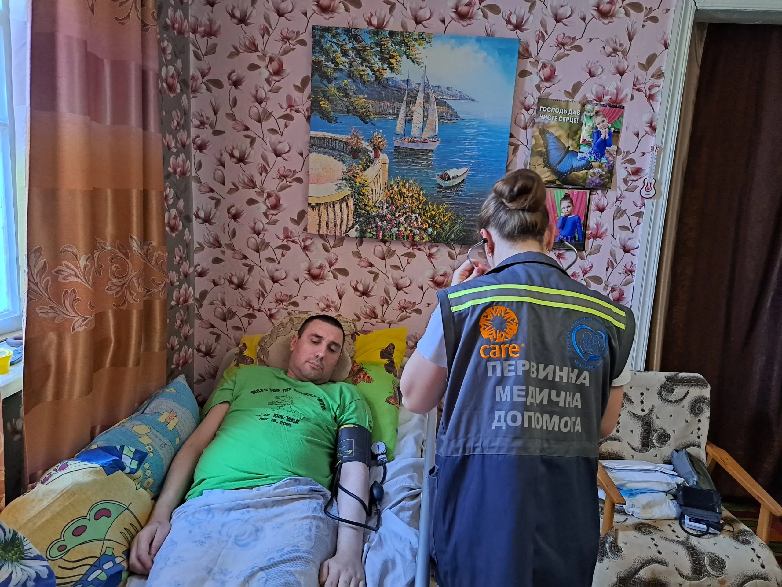 A man in a green shirt lays in a medical bed in Kramatorsk, Ukraine, as a medical professional in a CARE-branded vest performs an examination.