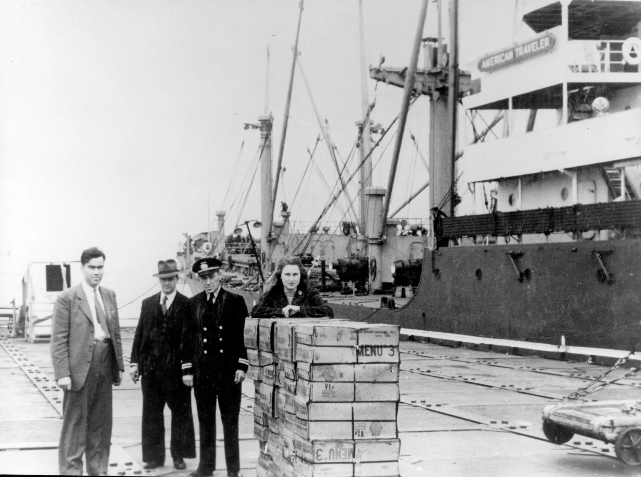 Four people stand in front of a large ship beside and behind a stack of original CARE PACKAGE parcels.