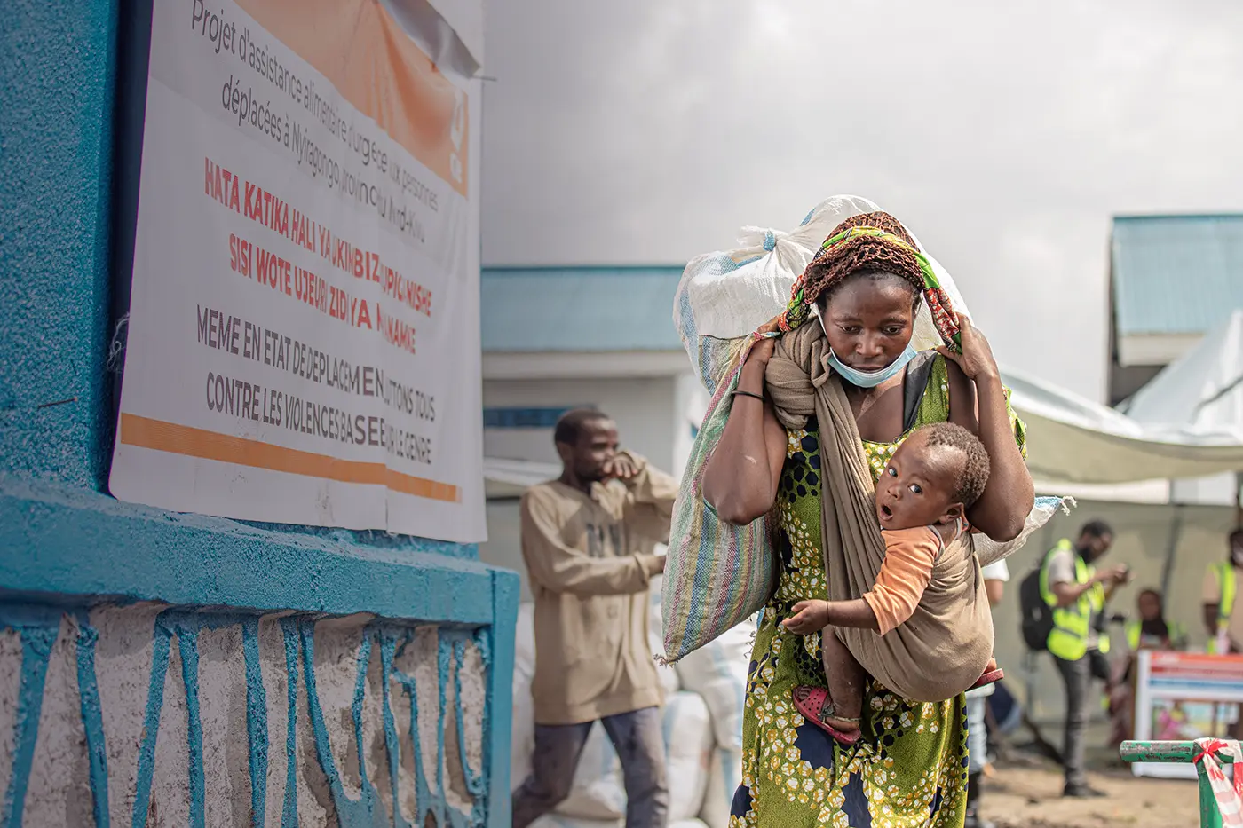 With a child strapped to her chest, a woman carries a heavy bag of food on her back.