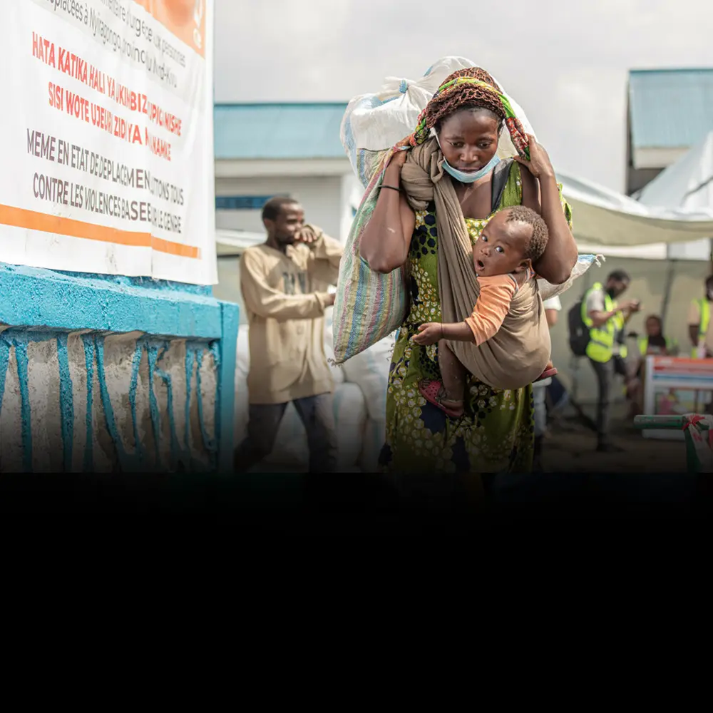 With a child strapped to her chest, a woman carries a heavy bag of food on her back.