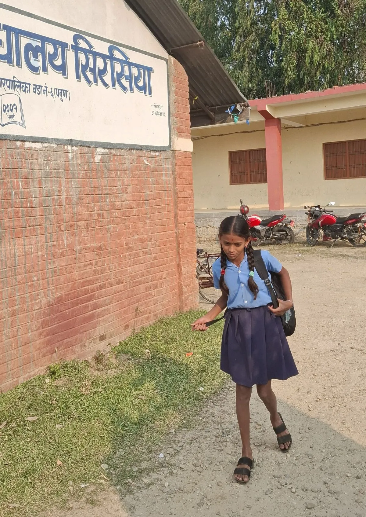 A young schoolgirl in a blue uniform and braids walks toward the entrance of Shree Secondary School, Sirsiya, carrying a backpack.