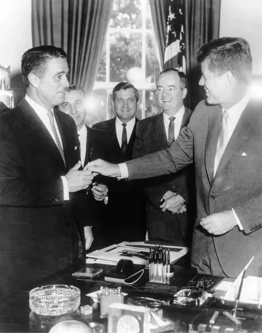 President John F. Kennedy stands with other men in suits in the Oval Office.