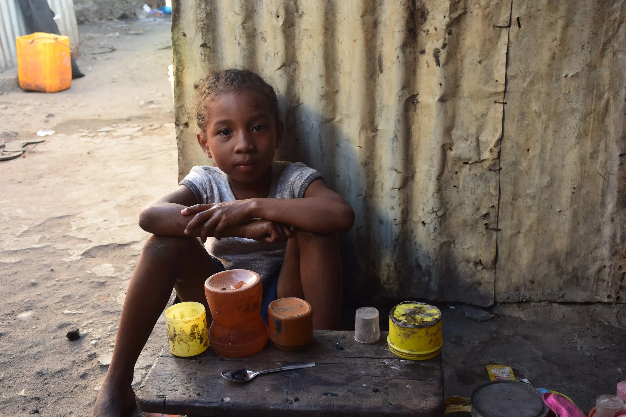 A child gazes at the camera while leaning against a corrugated iron (CI-sheet) wall.