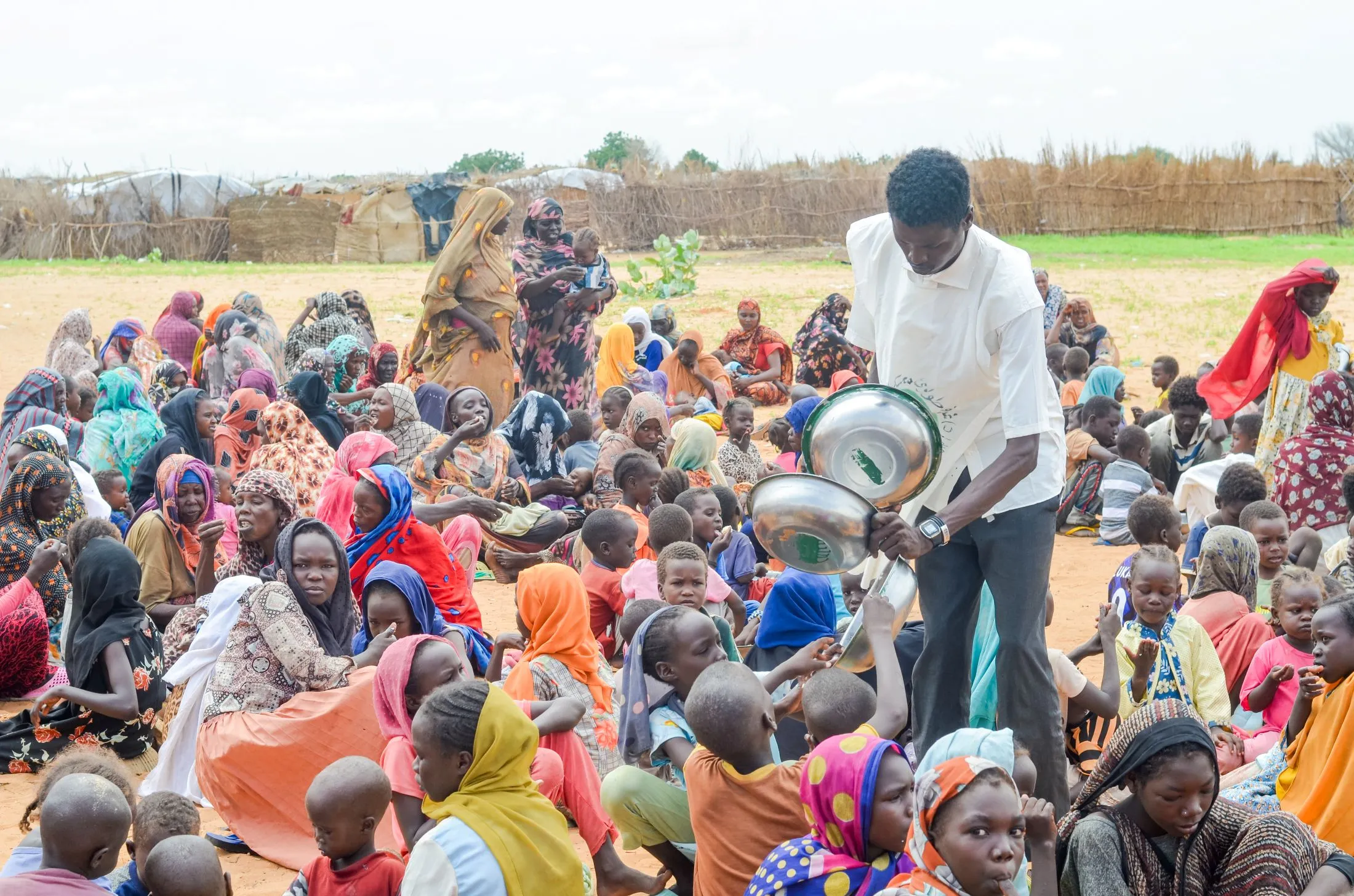 A man distributes food from metal bowls to a large crowd of Sudanese women and children. Many individuals in the densely packed group are wearing bright, patterned clothing.