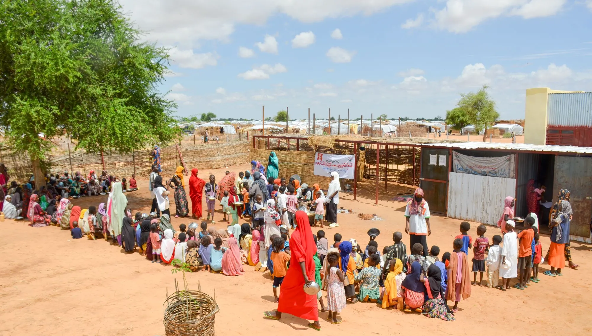A line of women and children, many in bright clothing, waits for assistance outside a distribution point in a Sudanese displacement camp. The scene shows temporary shelters and dry, sandy ground under a partly cloudy sky.