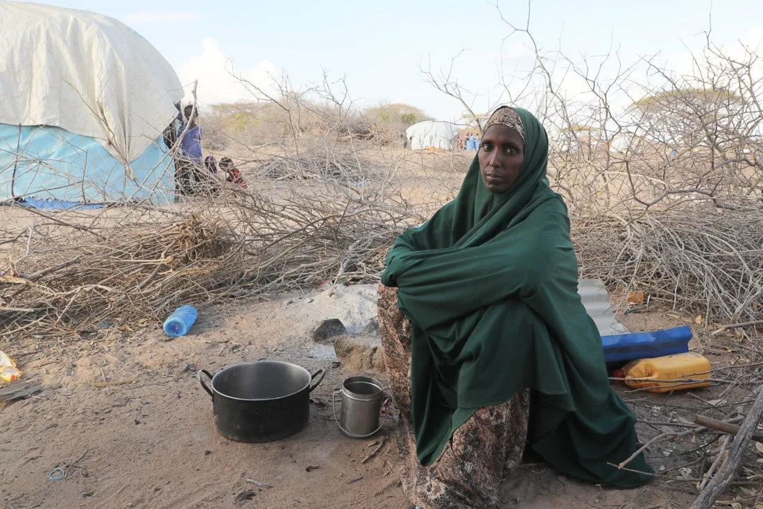 A Somali woman wearing a green shawl and patterned dress sits on the ground in a dry, sparse area surrounded by leafless branches, with an empty cooking pot and metal cup in front of her and dome-shaped tents in the background.
